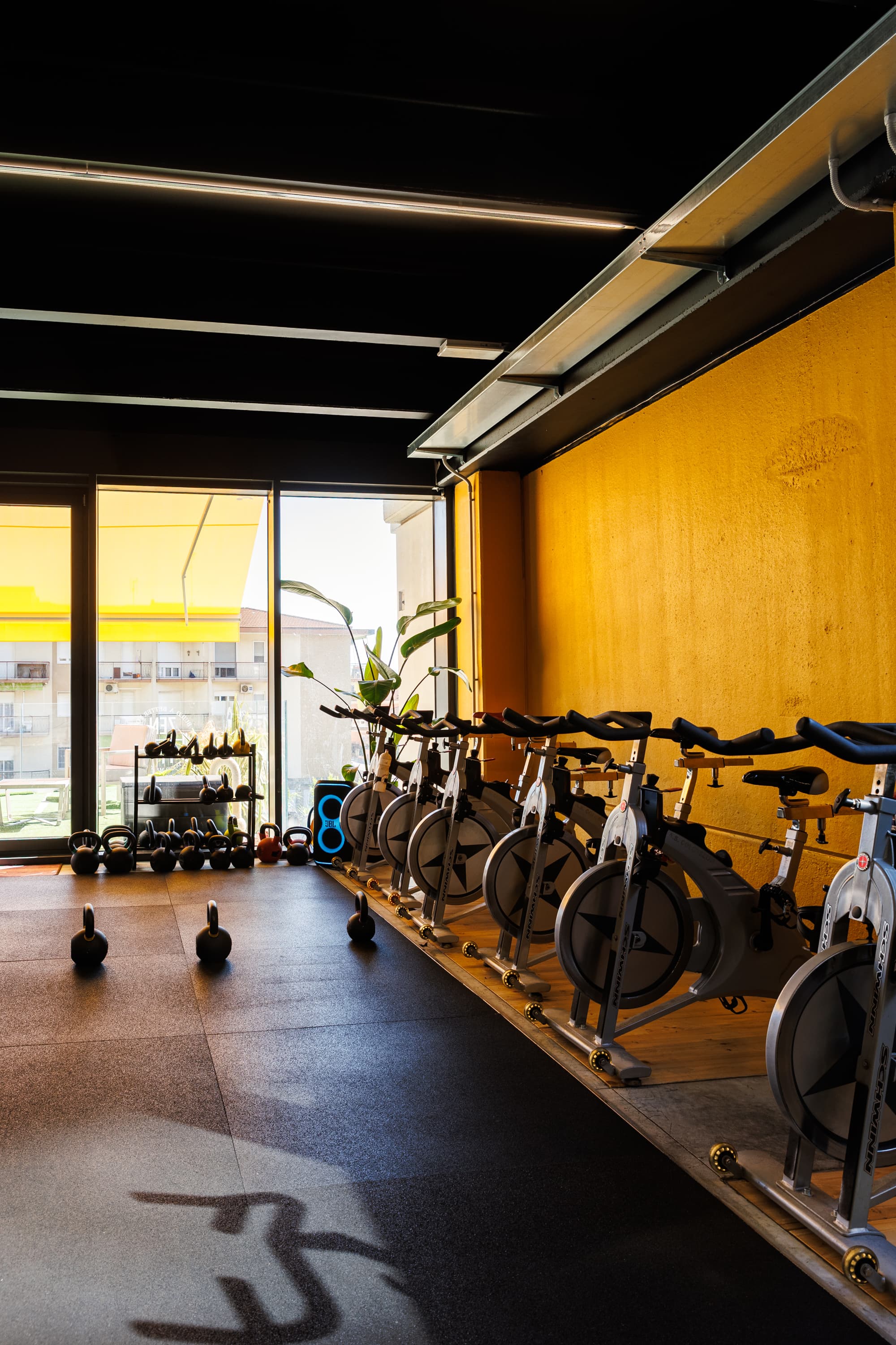 Le bike da spinning in fila sulla pedana, con i kettlebell a terra e la vetrata sulla strada.